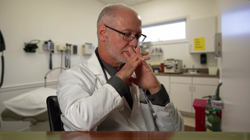 Male doctor wearing a lab coat and stethoscope in exam room sitting at desk dealing with depression or grief. Royalty-Free Stock Footage #1057590751