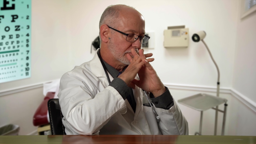 Male doctor wearing a lab coat and stethoscope in exam room sitting at desk dealing with depression or grief. Royalty-Free Stock Footage #1057590757