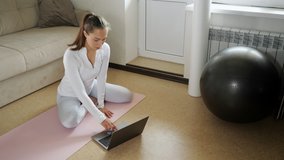 pretty woman with ponytail in white tracksuit surfs internet with modern laptop to find new exercises on mat near sofa in light room