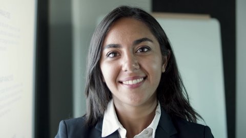 Closeup portrait of Latin beautiful young businesswoman standing in office room. Confident brunette secretary in suit smiling and looking at camera. Human resources, personnel and business concept