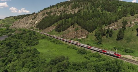 Passenger train an electric locomotive under the rock near river by two-sided winding Trans Siberian railway in the Ural Mountains - Aerial Photography, drone wide view at summer sunny day