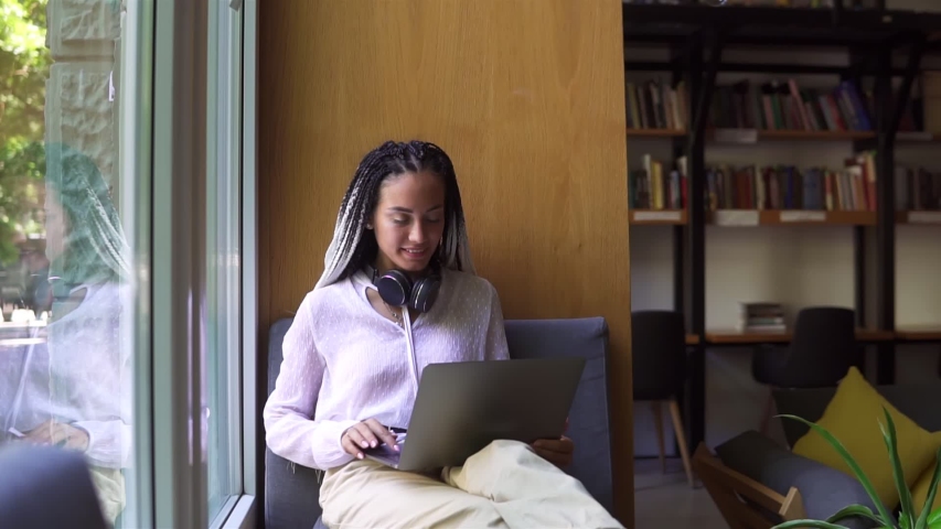 Stylish, dreadlocks student working on laptop at library or workplace. Young female professional working on computer device. Remote freelance work or studying - focused and concentrated. Wearing white Royalty-Free Stock Footage #1057595617