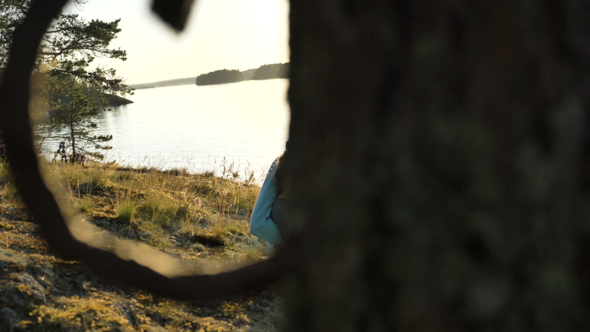 young woman and little girl child look at smartphone screen sitting on forestry bank of tranquil lake at bright sunset light
