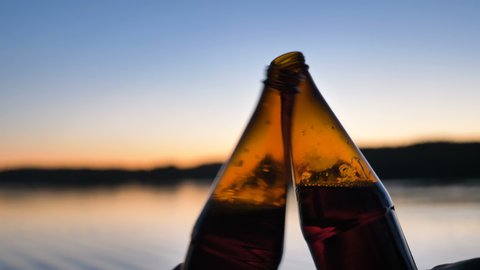 people clink brown glass bottles of beer against blurred view of large calm river and blue sky at sunset time close view