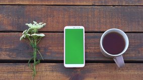 Closeup top view 4k video footage of white smartphone with empty blank green screen laying on wooden brown vintage background outdoors. Cute bouquet of meadow flowers laying near gadget, cup with tea.