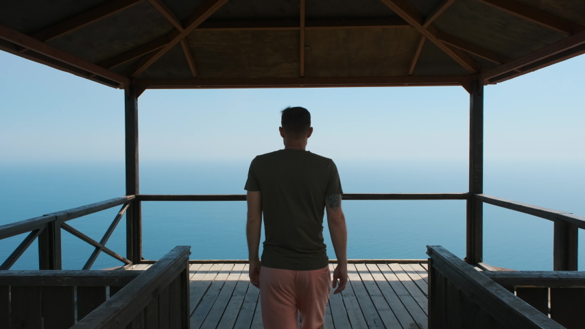 Young man on vacation comes to the railing, clouds on it and enjoys beautiful scenic view of the sea or ocean with ships in the distance. The guy looks at the ocean, follow up shot Royalty-Free Stock Footage #1057598548