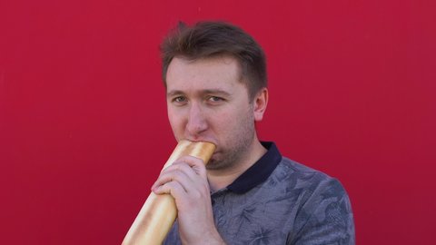 young man in a dark gray polo shirt, against background of red wall, on street chews and greedily bites off large piece of baguette, loaf with sad face, looks around, looks at camera, morning, day