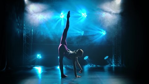 Rhythmic female gymnast performs acrobatic movements standing on hands. Slim gymnast in burgundy body performs the gymnastics exercise in a dark studio with neon lights. Silhouette. Slow motion.