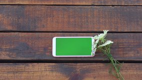 Closeup top view 4k video footage of modern smartphone laying on wooden brown vintage background outdoors. Cute small bouquet of wild meadow flowers laying near gadget with empty blank green screen.