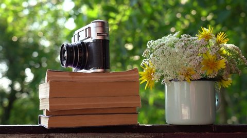 Closeup view 4k still life video footage of old vintage photo camera on stack of old paper books on brown wooden table background. Beautiful valley flowers arranged in blue enamel mug.