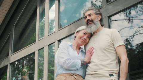 Low angle shot of happy senior woman standing on porch of vacation home with panoramic windows and talking to her husband. She is leaning her head on his chest and enjoying nature