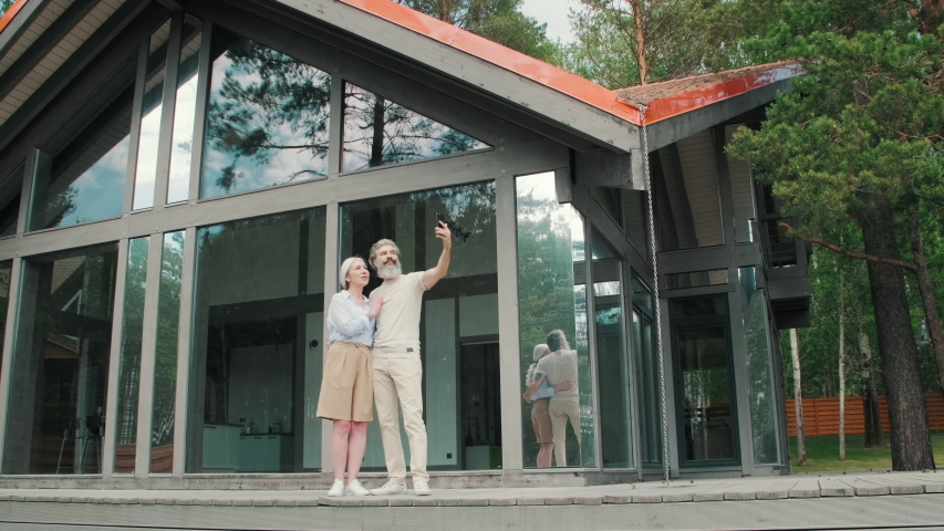 Lockdown shot of happy elderly woman and man with grey hair standing on porch of modern house with panoramic windows and taking selfie together