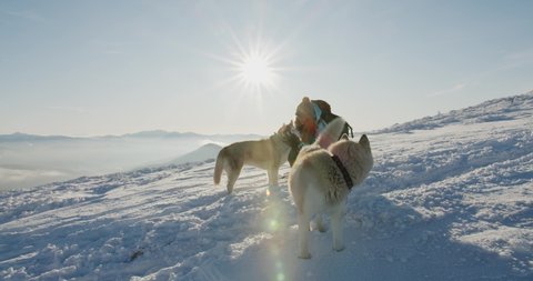 woman in blue jacket playing with two siberian husky dogs outdoors in snowy mountains. Hiker with dogs against beautiful winter mountain background. Active lifestyle and travel remote places concept
