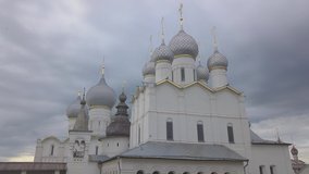White orthodox cathedral and churches with grey domes and golden elements against rainy cloudy skies with birds flying around. Low angle aerial shot slowly going up