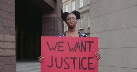 Afro american female student holding placard with we want justice writing on it. Female in glasses supporting human rights movement while standing at city street. Zoom in.