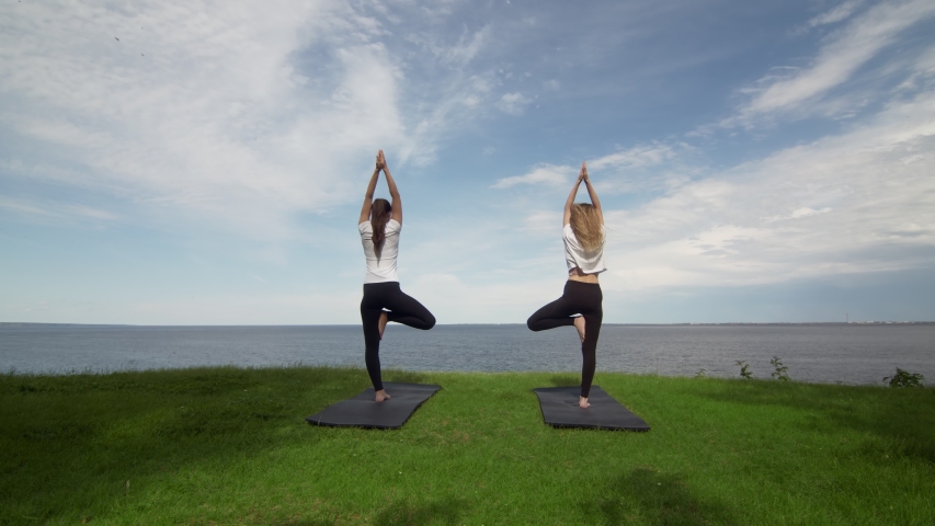 Two young women practice yoga on beach by the sea or ocea. Training tree pose.