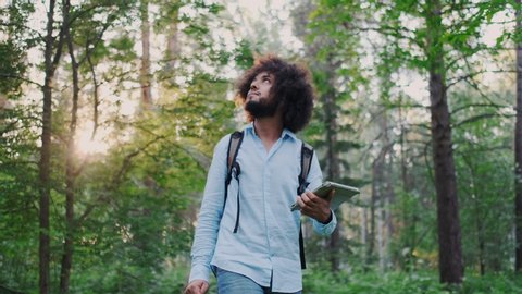 Young Curly Male Inspirational Environmentalist Walking in Forest. Nature Lover Hiking in Woods. Holding Tablet PC.Concept of Privacy and Isolation, Traveling Alone. Calmness, Harmony with Nature.