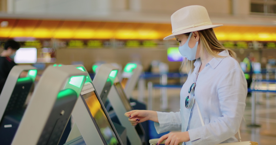 Woman in face mask checking-in at the airport. Safe travels under COVID-19. Caucasian attractive stylish traveler in coronavirus blue mask getting the boarding pass at station in empty terminal, 4K | Shutterstock HD Video #1057615843
