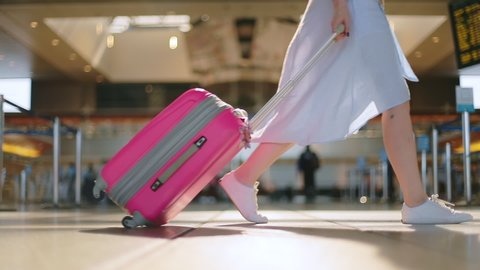 Close up view on traveler in the empty airport during Coronavirus outbreak. Slow motion of woman legs in white shoes and blue dress carrying her pink travel luggage on the wheels. Safe travels, USA