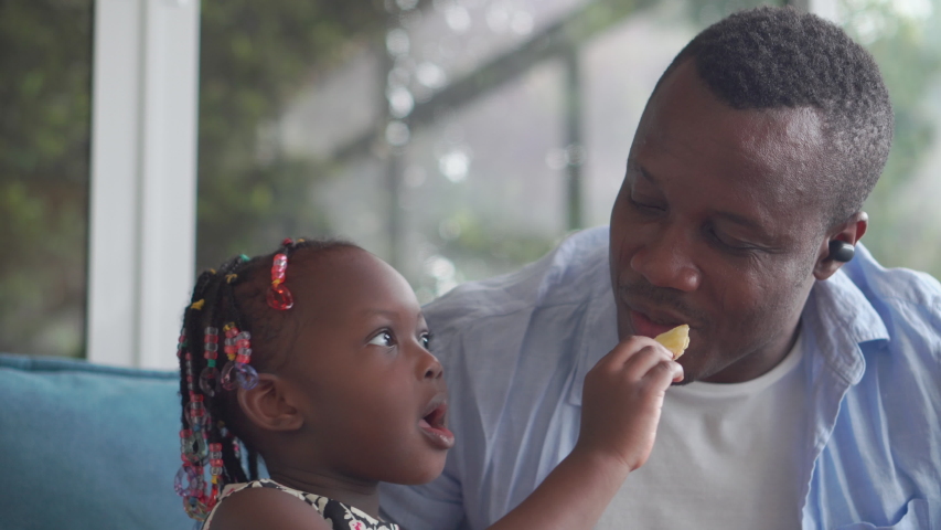 Cheerful african american father and daughter eating snack in living room, Happiness family concepts | Shutterstock HD Video #1057616650