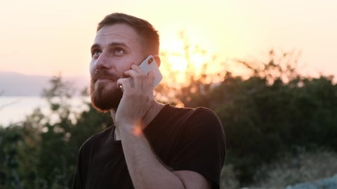 Bearded redhead is business man and constantly talks on the phone with his business partners. A nice guy is talking on the phone on vacation against the backdrop of a beautiful orange sunset.