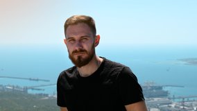 Confident guy in black T-shirt with red beard looks at camera. Video portrait of young man in the mountains against the background of the bay where the port is located.