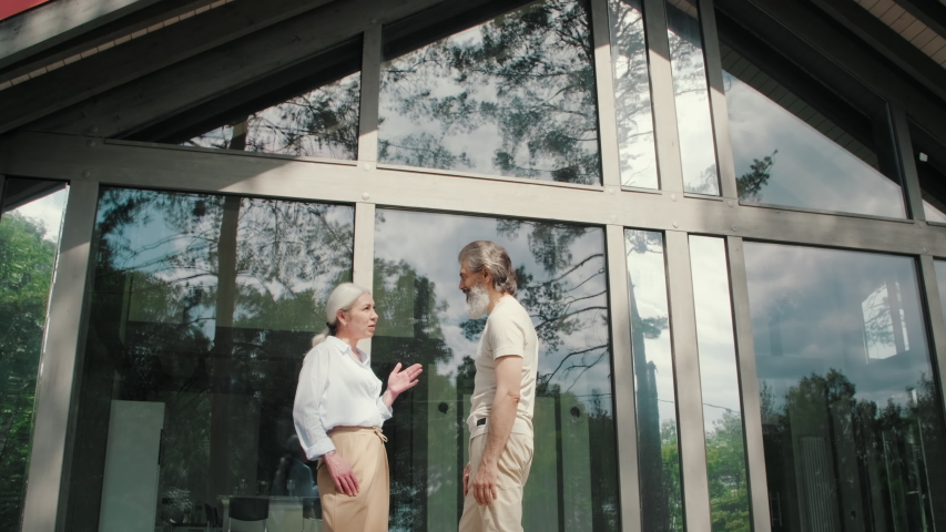 Tracking shot of wealthy senior couple with grey hair standing on porch of modern house with panoramic windows and having conversation