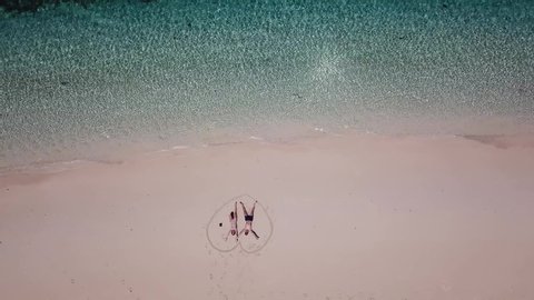 A drone shot of a couple lying on a pink sand beach in a heart drawn on the sand a small island near Maumere, Indonesia. Happy and careless moments. Waves gently washing the shore. Romance and love
