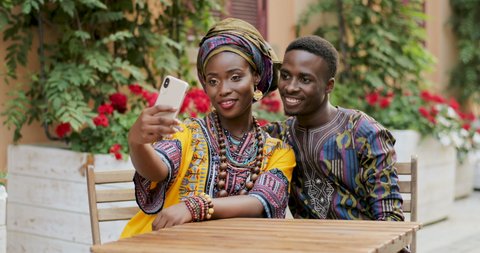 Beautiful young African woman and attractive man sitting at the table in the yard with flowers and smiling to the smartphone camera while taking selfie photos.