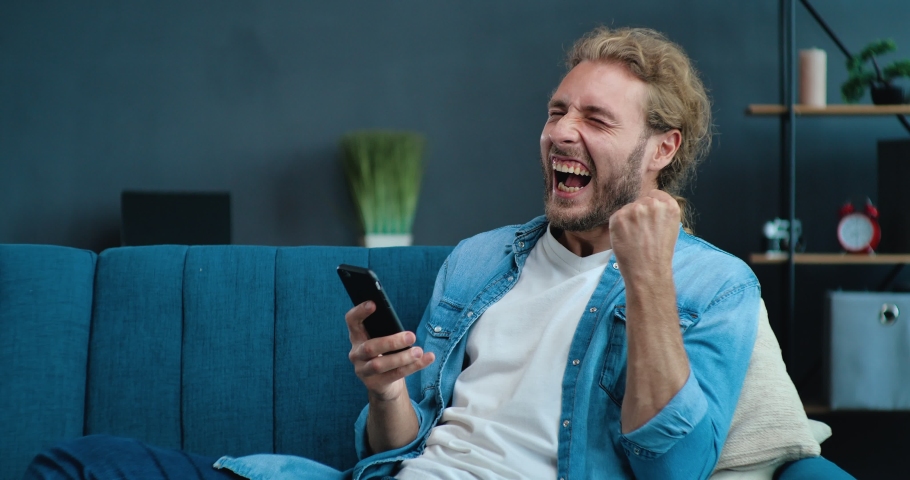  Happy Man celebrating mobile win uses Phone at home office. Man reading good news, Victory while sitting on couch | Shutterstock HD Video #1057627729