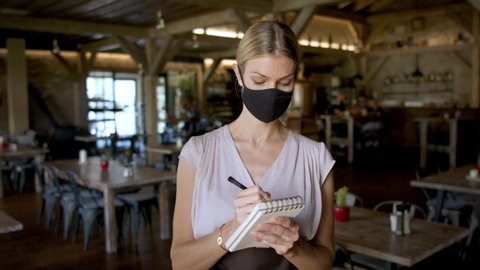 Waitress with face mask and order pad standing indoors in restaurant.