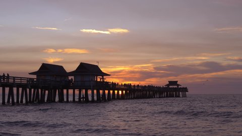 SLOW MOTION: Naples Beach and Fishing Pier at Sunset, Florida. Dark silhouette of a pier over the water at sunset. Sunset over horizon and wooden pier of caribbean beach of Naples in Florida. Sunset.