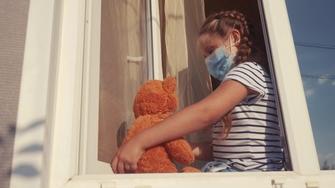 bored kid in medical mask in home quarantine covid 19 coronavirus sitting by the window. child with a toy teddy bear in protective mask looking out the window. coronavirus epidemic prevention concept