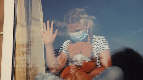 bored kid in medical mask in home quarantine coronavirus sitting by the window. child with a toy teddy bear in protective covid 19 mask looking out the window. coronavirus epidemic prevention concept