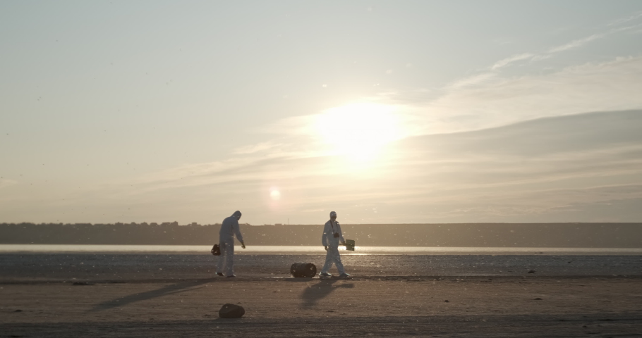 Two scientists in white suits walk around a black barrel that lies on the coast. Biological inspection of objects polluting the environment. | Shutterstock HD Video #1057639078
