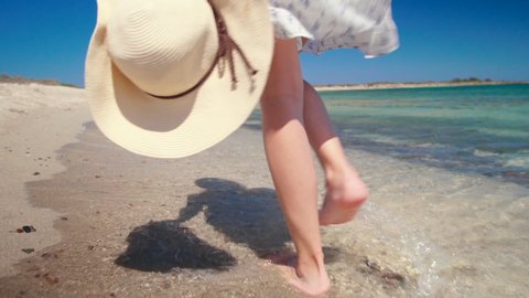Female Foot Steps on Beach. Barefoot Woman Walks along Sea Coast with Hat in Summer Sunny Day. Holiday and Vacation on Chrysi, Crete, Greece. 4K Handheld Close up Tracking Backside Shot