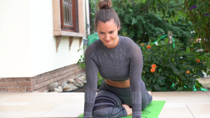 Beautiful young woman watching video on laptop and practicing yoga pose in backyard. High quality 4k footage