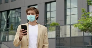 Handsome man in formal clothing and medical mask using modern smartphone while walking on street. Concept of pandemic, work and technology.