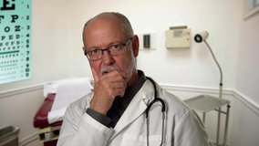 Portrait of experienced gray haired doctor with glasses in exam room listening to someone, thinking and indicating motion in the shoulder.