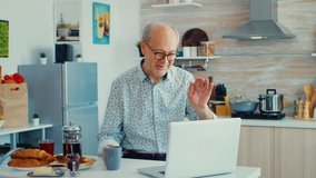Senior man chatting and waving during a video call using laptop in kitchen holding a cup of coffee. An elderly person using internet online chat technology video webcam making a video call connection. - Powered by Shutterstock - Get 15% off with code: PIKWIZARD15