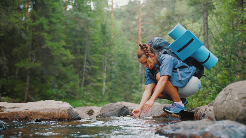 Woman traveler drinks clean water from river or spring in summer forest. Female tourist with backpack drinking fresh mountain water while a hiking, scooped up cold transparent water with his hands. Royalty-Free Stock Footage #1057659466