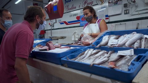 DENIA, SPAIN – AUGUST 11 2020. A fishmonger wearing a face mask cleans fresh fish at Denia food market as a male customer wearing a face mask awaits in the new normal after Cronavirus Covid pandemic
