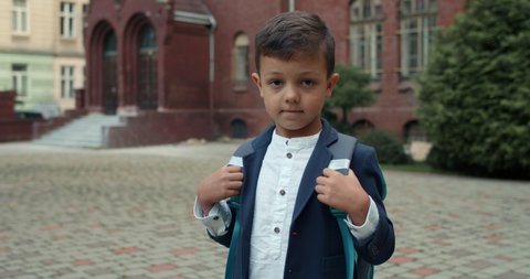 Portrait of small boy with backpack looking to camera. Crop view of kid in school uniform posing while standing at street. Concept of education and children. 