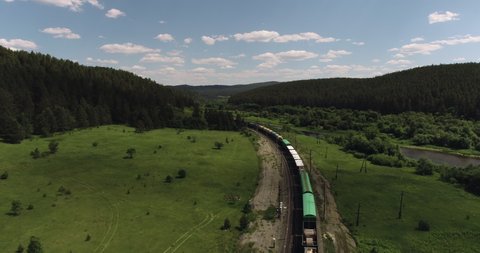 Freight long train carries with cargo carriages in a village in wild mountains landscape through a difficult part of Trans Siberian railways. Aerial drone wide view at summer day.