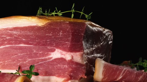 large piece of prosciutto and chopped pieces rotate on a wooden board around its axis, black background