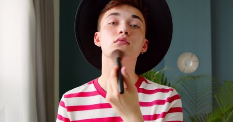 Young transgenger queer man make up beauty routine standing in front of camera. A metrosexual is applying powder with a brush, preparing for a date. 