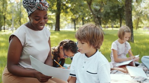 Smiling Afro-American female teacher smiling and explaining task in workbook to little Caucasian boy during outdoor lesson in the park