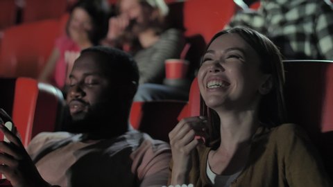 Close-up portrait of beautiful asian woman and handsome bearded dark-skinned man eating popcorn and laughing during funny movie. Multi-ethnic viewers having fun while watching comedy in cinema theater