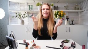 Vlogger female applies lipstick on lips. Beauty blogger woman filming daily makeup routine tutorial at camera on tripod. Influencer blonde lady live streaming cosmetics product comparison in studio