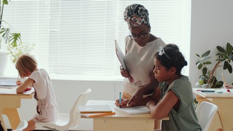 Afro-American female teacher walking through classroom and helping children while they writing in notepads, then standing beside flipchart and explaining something during lesson